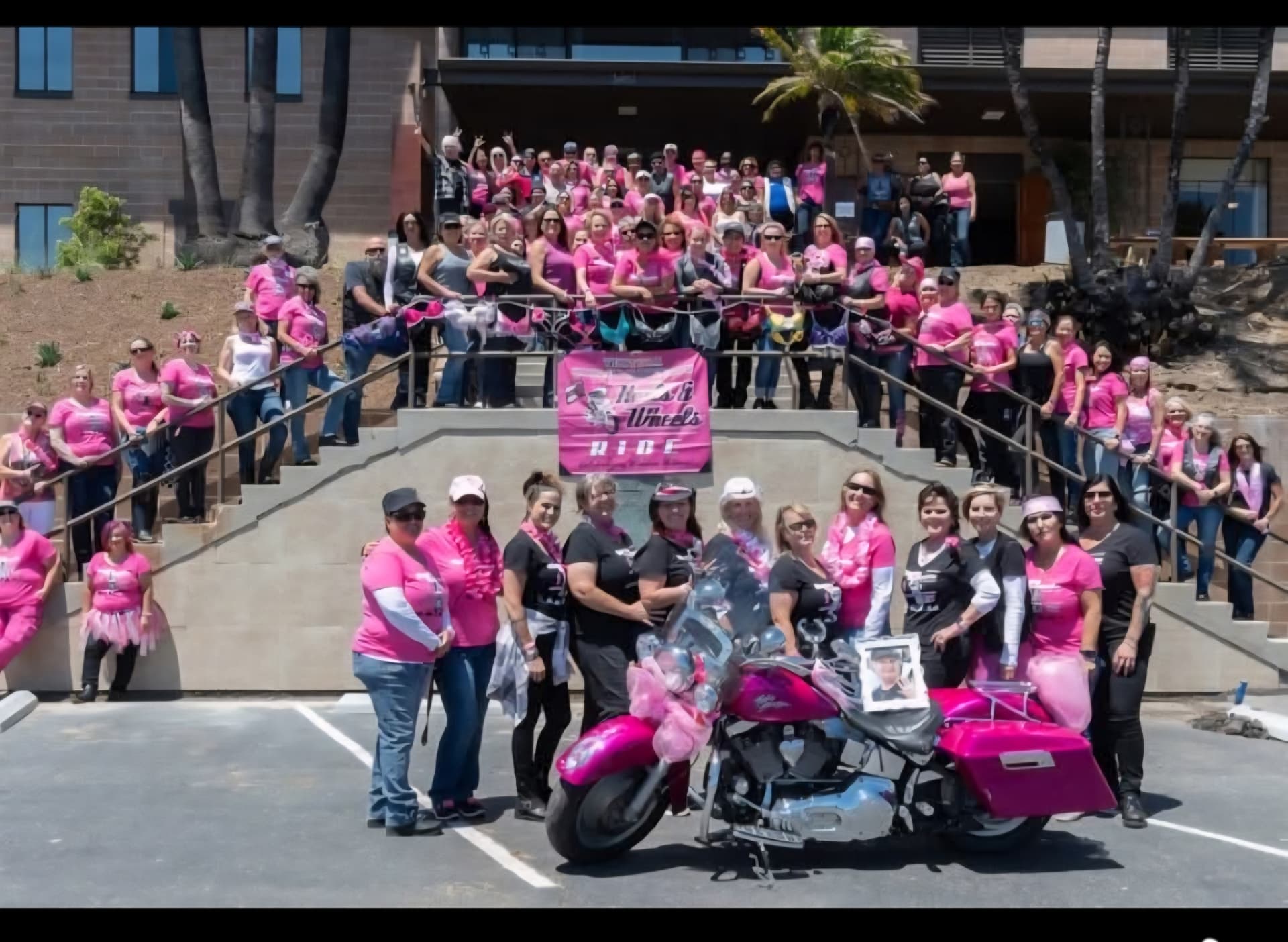Large group of Heels & Wheels riders gathered on the steps behind a pink Harley-Davidson