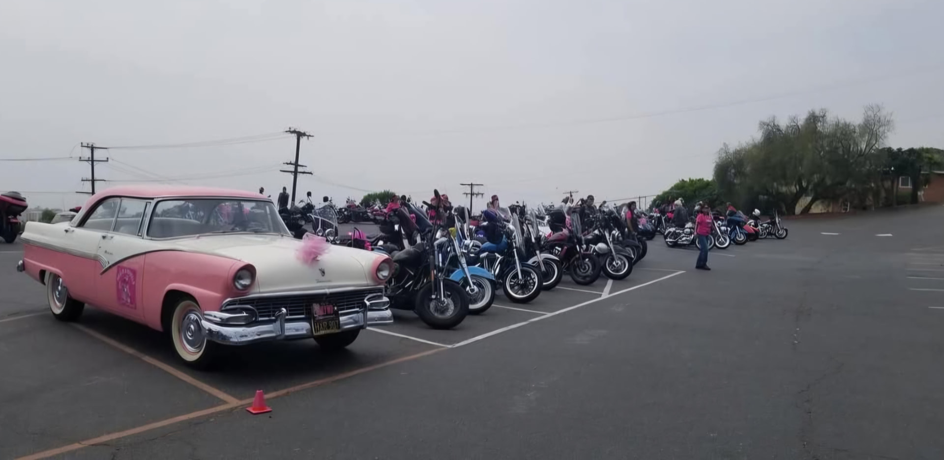 Pink and white classic car parked alongside rows of motorcycles at the ride staging lot